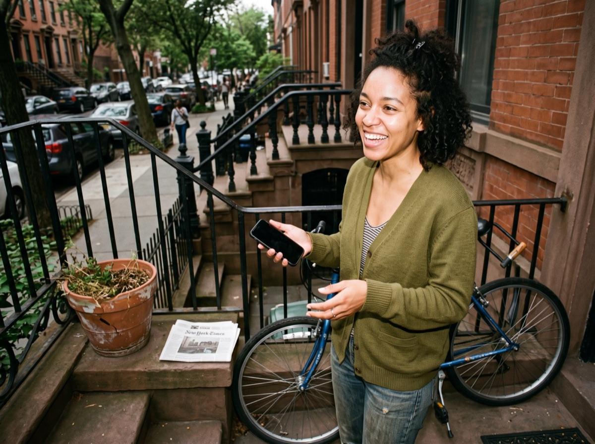 A woman on her front stoop, phone in hand, laughing mid-conversation beside her bike